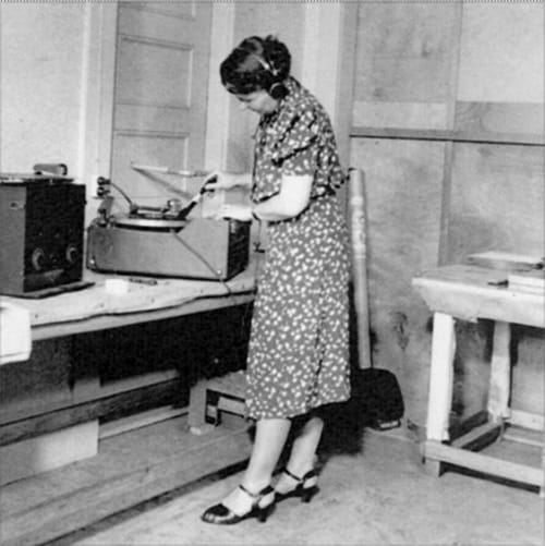 A woman in a floral dress skillfully operates a vintage recording device on a table, enhancing the sophisticated and high-value ambiance of the room.