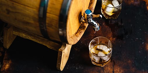 A premium whisky barrel with a tap sits beside two glasses of iced, top-shelf brown liquid on a rich, dark wooden surface.