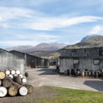 Nestled in a distillery yard, the stacked barrels and grey buildings are framed by the majestic backdrop of Ben Nevis, creating a scenic and iconic setting under a partly cloudy sky—ideal for showcasing high-value whisky investment opportunities.