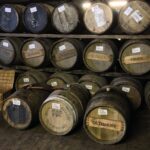 In a dimly lit storage area, rows of premium wooden barrels are stacked neatly, each adorned with distinctive labels and markings, reflecting the high-value whisky investments stored within.