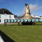 A prominent distillation apparatus stands impressively outside the pristine white building, decorated with blue circular markers inscribed with "Bruichladdich." This iconic scene invites whisky enthusiasts and investors, symbolizing lucrative Bruichladdich cask whisky investment opportunities.