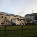 The industrial building, with its commanding smokestack echoing the sturdy elegance of a Clynelish cask, stands gracefully amid lush grass and a rustic wooden fence. This scene is set beneath an evocative, cloudy sky, representing the high value and quality often associated with whisky investments.