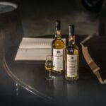 Two bottles of high-value Clynelish cask whisky alongside a filled glass sit elegantly on a wooden table, framed by an investment notebook and a curved tasting tool—a quintessential setting for discerning whisky investors exploring quality opportunities.