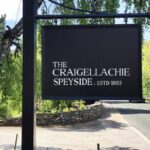 A sign displaying "The Craigellachie Speyside. Estd 1893" stands against a scenic backdrop of lush trees and a winding road, encapsulating the timeless elegance and premium investment potential of Craigellachie's renowned cask whisky heritage.