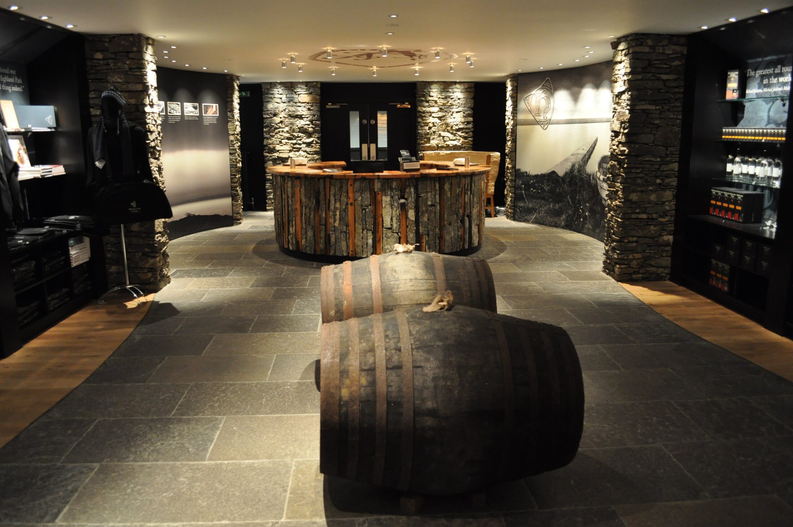 A sophisticated whiskey tasting room featuring stone walls and wooden barrel decor seamlessly blends luxury with tradition. This high-value space includes merchandise displays and a central counter, embodying quality and elegance for discerning investors in the whiskey market.