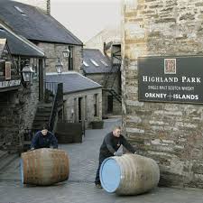 At a prestigious distillery, showcasing high value whisky investments, two individuals roll large barrels along a cobblestone path beneath a sign on the brick wall that reads "Highland Park, Single Malt Scotch Whisky, Orkney Islands," emphasizing both quality and tradition.