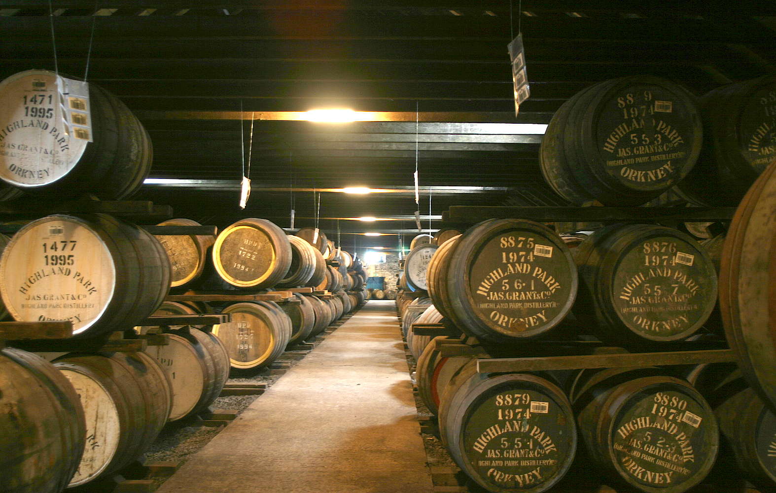 A dimly lit warehouse showcases rows of stacked wooden barrels, each intricately labeled with prestigious whisky brands like "Highland Park" and detailed dates, embodying investment potential and quality craftsmanship.