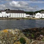 Discover the allure of Laphroaig Cask Whisky Investment with the iconic white buildings of the Laphroaig distillery, nestled by the coastline. Set against a dramatic partly cloudy sky, with a striking rocky foreground and shimmering water in front, this picturesque scene captures the essence of premium whisky investing.
