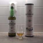 The showcase features a bottle of Laphroaig 10 Year Old Islay Single Malt Scotch Whisky, ideal for whisky investment enthusiasts. Alongside sits a tasting glass filled with the premium whisky and its original packaging, all elegantly arranged on a countertop against a sophisticated tile backsplash.