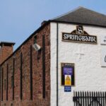 This image captures the iconic exterior of Springbank Distillery, featuring its distinctive brick and white walls adorned with signage for prestigious whisky brands like Springbank, Longrow, and Campbeltown Malts. Set against a backdrop of a clear blue sky, it embodies the quality and allure of high-value whisky investments.