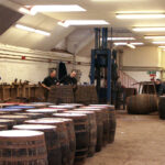In a premium whisky investing warehouse, wooden barrels are meticulously stacked high, surrounded by three skilled workers. The room showcases pristine white walls and a lofty ceiling illuminated by bright overhead lighting, reflecting the quality of our investments.