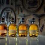 Four high-value whisky investment bottles are elegantly arranged on a pristine white surface, set against a dark blurred background featuring sophisticated tools.