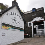 Explore lucrative Tobermory Cask Whisky investment opportunities with an exterior view of the historic Tobermory Distillery, established in 1798. This iconic site features buildings adorned with signage for Island Single Malt Scotch Whisky, epitomizing high-value investments and rich heritage.