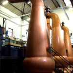 Inside the industrial facility, gleaming copper distillation stills are surrounded by intricate metal walkways and pipes, underscoring the quality craftsmanship associated with high-value whisky investments.