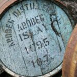 A weathered wooden barrel, adorned with faded blue paint and black text, dated 1995, suggests an Ardbeg cask—a symbol of high-value whisky investment. Resting against the wall, it whispers tales of premium spirits.