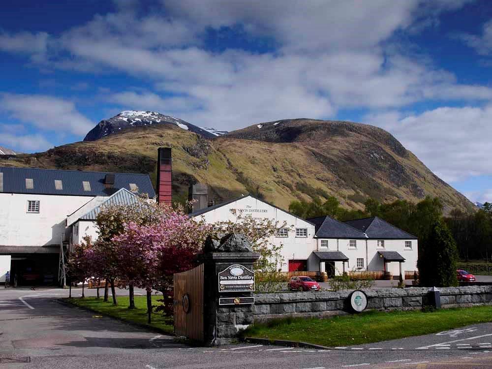 The distillery buildings framed by the majestic Ben Nevis stand out against a partly cloudy sky, embodying the essence of high-value whisky investment. This picturesque blend of iconic Scottish landscapes and premium whisky heritage offers a visual testament to quality and sophistication.