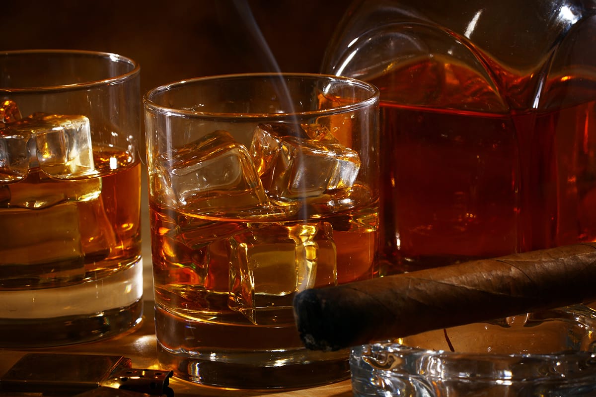 Two glasses of premium whisky, filled with rich amber liquid and ice cubes, sit elegantly beside a fine cigar, while a luxurious decanter graces the background—a perfect tableau of high-value investments and quality craftsmanship.