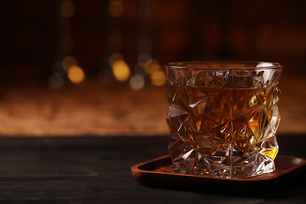 A premium faceted glass tumbler, brimming with rich amber whisky, rests elegantly on a sleek wooden investment tray, set against a sophisticated dark backdrop.