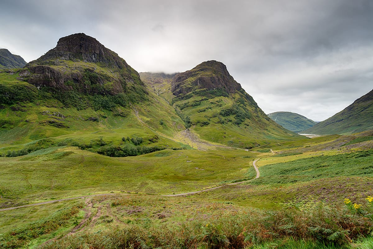 Explore a breathtaking vista of a lush green valley, framed by two majestic hills, under a dramatic cloudy sky. This picturesque landscape features a winding path that invites wanderers and embodies the premium quality and sophisticated nature synonymous with high-value whisky investments.