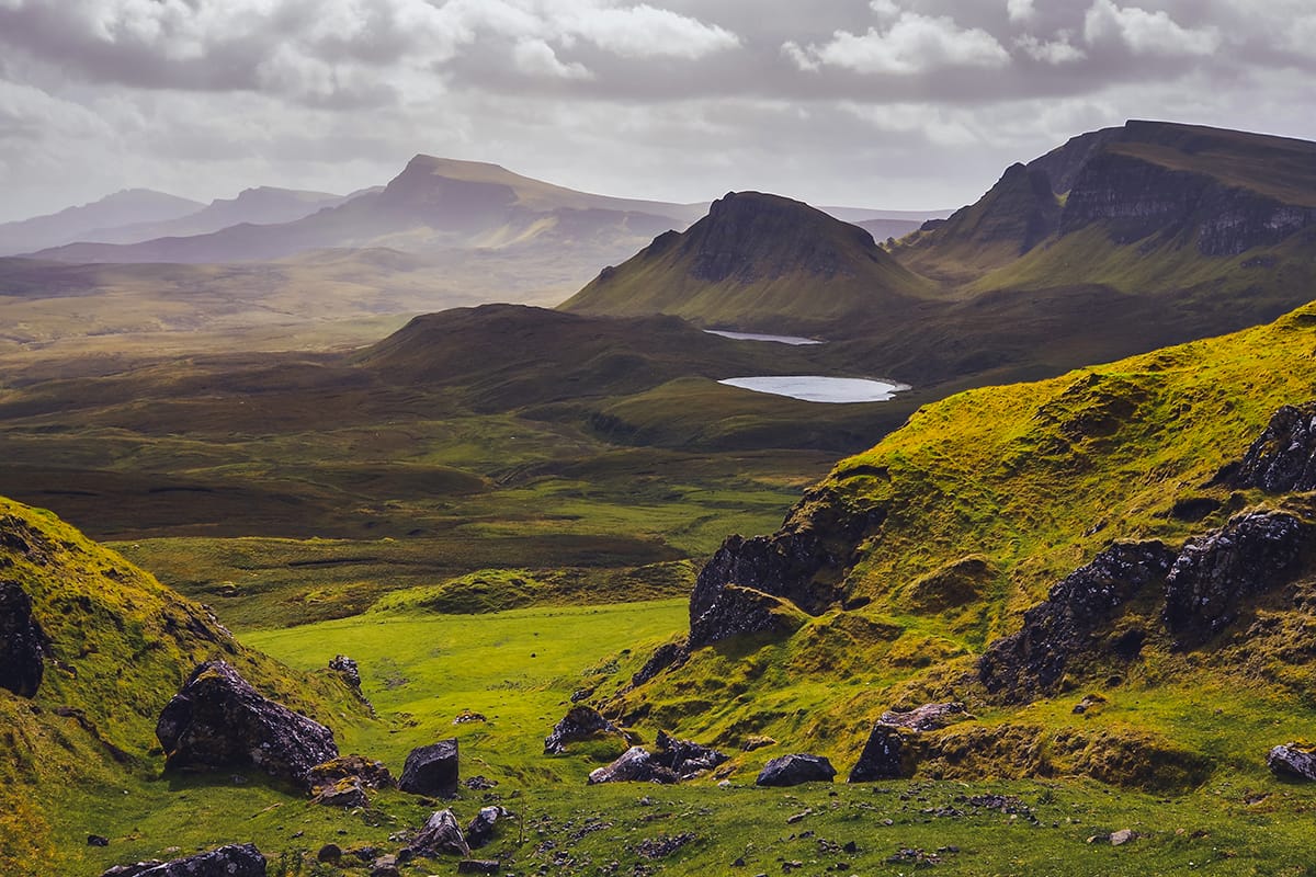 Rolling green hills with rocky outcrops and a small lake under a cloudy sky create a picturesque landscape, reflecting the quality and elegance of high-value whisky investments. Distant mountain peaks add to the breathtaking backdrop, symbolizing the maturity and timeless appeal of premium whisky brands.