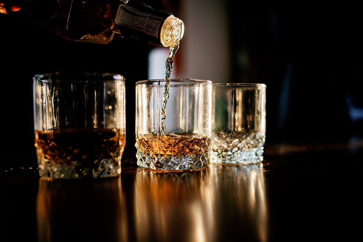 A premium whiskey flows elegantly into one of three exquisite crystal glasses, strategically placed on a reflective surface. The dark background enhances the luxury and sophistication, underscoring its high-end appeal and investment value.