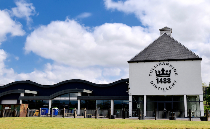This captivating image showcases the Tullibardine Distillery exterior, featuring a pristine white building with a distinctive wavy roof set against a vibrant blue sky dotted with clouds. Perfectly embodying high-value whisky investing, this visual highlights both the elegance of premium brands and the quality often sought in investment opportunities.