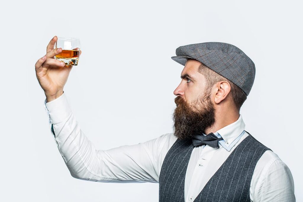 A bearded man, sporting a flat cap and waistcoat, thoughtfully examines a glass of premium amber liquid against a light backdrop, reflecting on the intricacies of whisky investment and high-value brands.