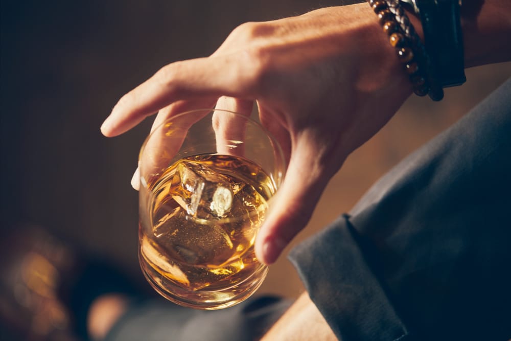 A whisky investor holds a glass of premium Scotch whisky on the rocks, showcasing their bracelet and dark trousers—an elegant scene that captures the sophistication of whisky investment.