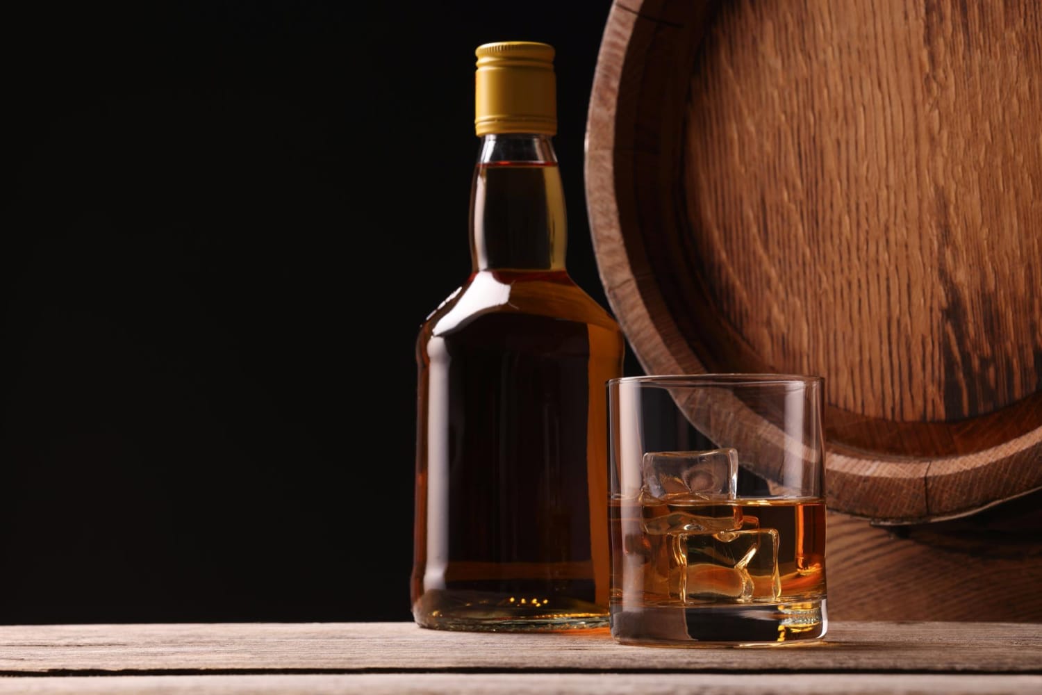 A premium bottle of whisky, a glass filled with whisky and ice cubes, and an aged oak barrel sit on a rustic wooden surface, creating an elegant display ideal for any whisky investment website.