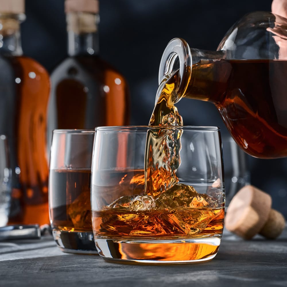 A crystal decanter pours premium whisky into a glass with ice, with another whisky glass and investment bottles in the background.