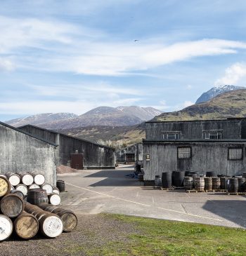 Nestled in a distillery yard, the stacked barrels and grey buildings are framed by the majestic backdrop of Ben Nevis, creating a scenic and iconic setting under a partly cloudy sky—ideal for showcasing high-value whisky investment opportunities.