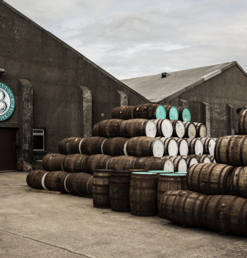 Under a dramatic, cloudy sky, the Bruichladdich warehouse exterior proudly displays its iconic logo. The sight of stacked wooden barrels hints at exclusive cask whisky investment opportunities and the potential of high-value brands within its storied walls.