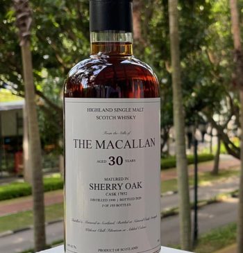 A rare and exquisite bottle of The Macallan 30 Years Old Highland Single Malt Scotch Whisky, aged in sherry oak, is displayed elegantly on a ledge surrounded by lush greenery, depicting both luxury and nature's beauty.