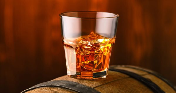A glass of premium, investment-grade whisky with ice sits atop a wooden barrel, set against a warm, blurred background that evokes quality and value.