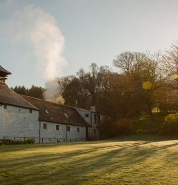 The historic Glengoyne distillery building stands elegantly under a clear sky, enveloped by lush trees and grassy fields. Sunlight illuminates the scene, inviting whisky investors to explore premium cask whisky investment opportunities nestled within its storied walls.